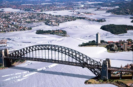 The Sydney Harbour Bridge is in the foreground and the islands and foreshores of the western harbour are in the middle distance. The city stretches out to the west in the background. The water of the harbour is white with gleaming ice like the surface of Antarctica.